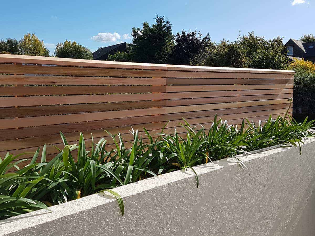 A cedar slatted fence positioned in a flower bed on top of a wall.