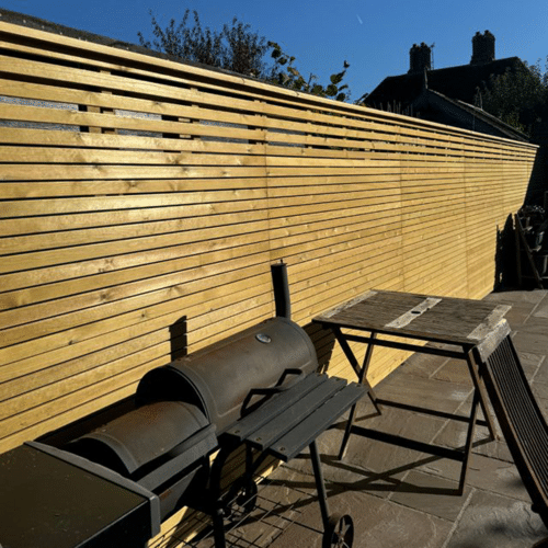 A horizontal slat fence in a sunny garden with a patio and barbeque.